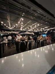 Rooftop patio at night with hanging string lights and a group of people in aprons gathered around long high tables for a communal cooking class/outdoor dining event