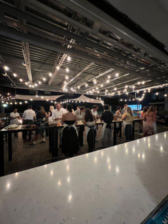 Rooftop patio at night with hanging string lights and a group of people in aprons gathered around long high tables for a communal cooking class/outdoor dining event