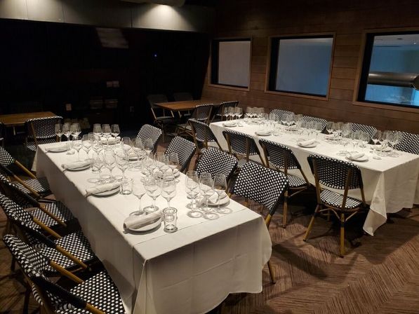 Cozy wood-paneled private dining room in a restaurant with two long white-tablecloth tables set for a group wine tasting or dinner, lined with multiple wine glasses and black-and-white bistro chairs.