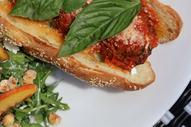 Italian meatball sandwich on a sesame-crusted baguette with marinara, grated Parmesan and fresh basil, served with arugula salad, peach slices and hazelnuts on a white plate