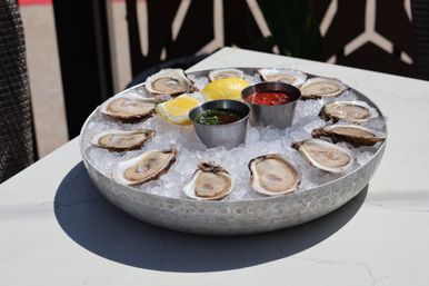 Fresh oysters on the half shell arranged on crushed ice with lemon wedges and metal cups of cocktail sauce and mignonette on a sunlit patio table