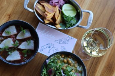 Top-down view of a wooden table with citrus-marinated sliced fish (ceviche), chickpea and greens salad with radish, plantain chips with lime and pickled onions, and a glass of white wine.