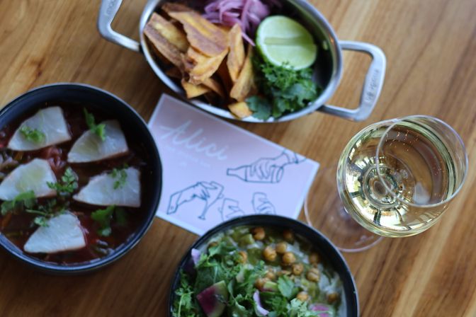Top-down view of a wooden table with citrus-marinated sliced fish (ceviche), chickpea and greens salad with radish, plantain chips with lime and pickled onions, and a glass of white wine.