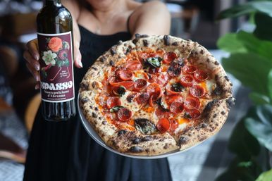 Neapolitan-style wood-fired pepperoni pizza with blistered crust, melted mozzarella and basil, held alongside a bottle of red wine in a casual restaurant setting.