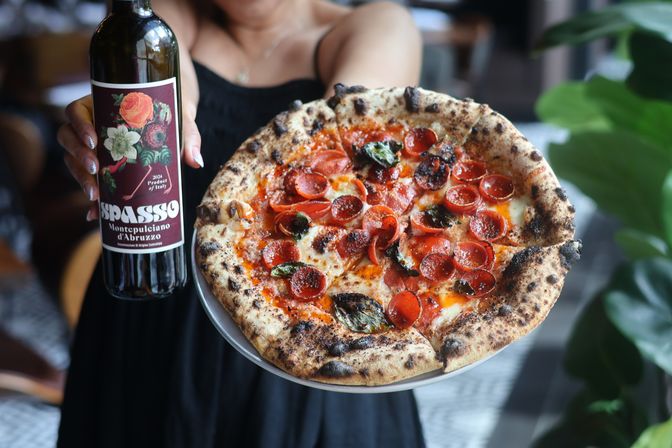 Neapolitan-style wood-fired pepperoni pizza with blistered crust, melted mozzarella and basil, held alongside a bottle of red wine in a casual restaurant setting.