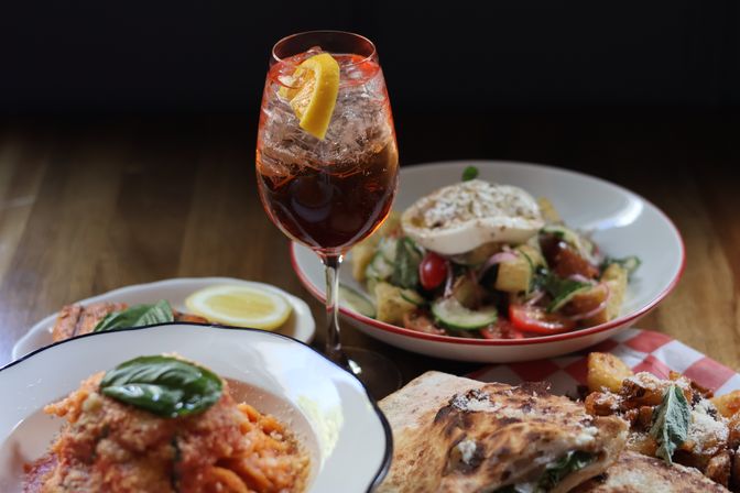 Italian-style spread on a wooden table: tall orange spritz garnished with a lemon twist, bowl of tomato pasta with basil, panzanella salad with burrata, and wood-fired pizza with roasted potatoes.