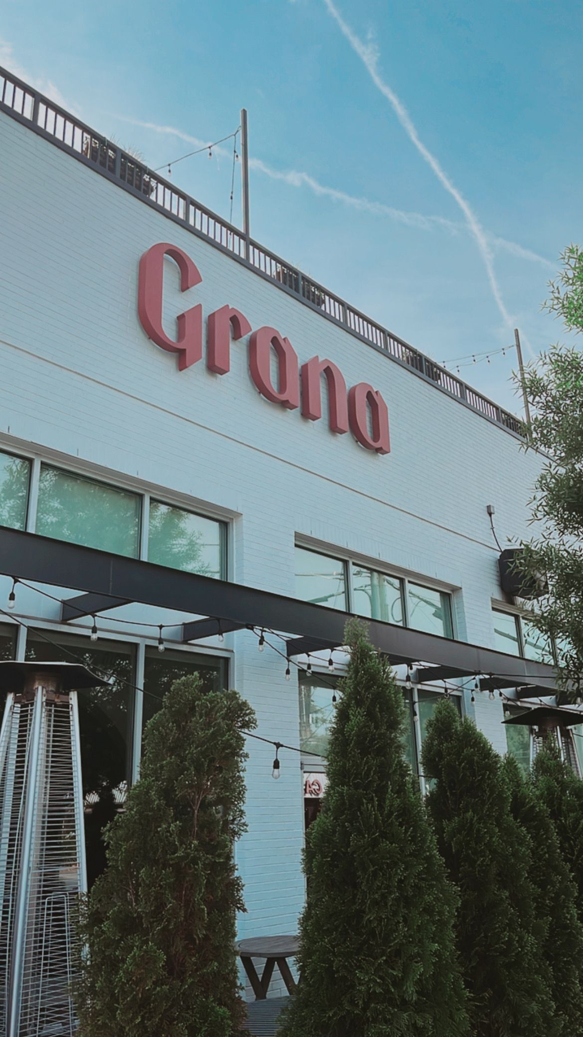 White brick urban cafe facade with large red signage, rooftop railing and string lights against a clear blue sky; outdoor patio framed by tall evergreen shrubs, windows and patio heaters.