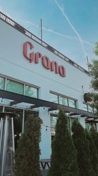 White brick urban cafe facade with large red signage, rooftop railing and string lights against a clear blue sky; outdoor patio framed by tall evergreen shrubs, windows and patio heaters.
