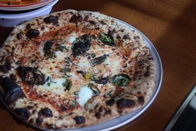 Wood-fired Neapolitan pizza with charred blistered crust, melted mozzarella, fresh basil, and tomato sauce on a metal pan atop a wooden table.