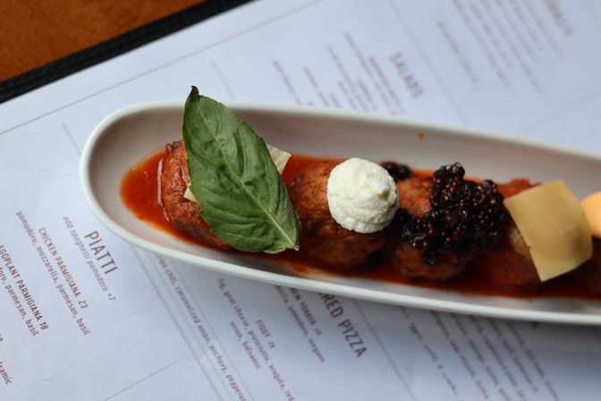 Gourmet Italian-style meatballs in tomato sauce on a long white plate, topped with whipped ricotta, a large basil leaf and shaved Parmesan, photographed over a restaurant menu.