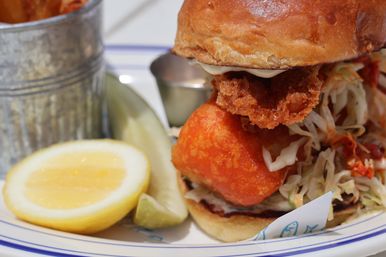 Close-up of a crispy golden battered fish sandwich on a brioche bun with tangy coleslaw, lemon wedge, pickle spear and a metal cup of fries, plated seafood dish.