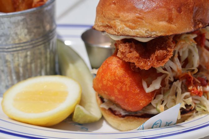 Close-up of a crispy golden battered fish sandwich on a brioche bun with tangy coleslaw, lemon wedge, pickle spear and a metal cup of fries, plated seafood dish.