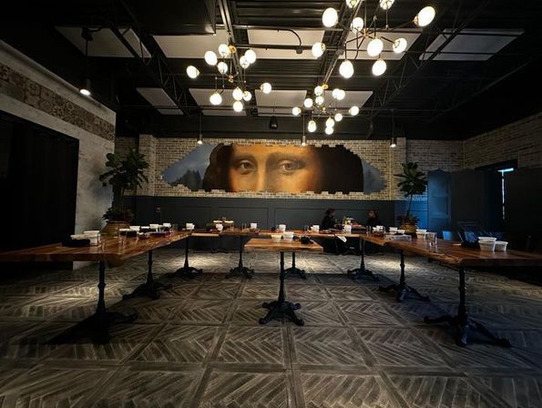 Industrial restaurant interior with long wooden communal tables, patterned tile floor, globe chandeliers, exposed brick walls and a large classical portrait mural revealed through a faux broken-brick wall.