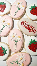 Assorted hand-iced sugar cookies with pastel pink and white icing, piped daisy flowers, red strawberry designs and one cookie with red script lettering — bright spring-themed treats.