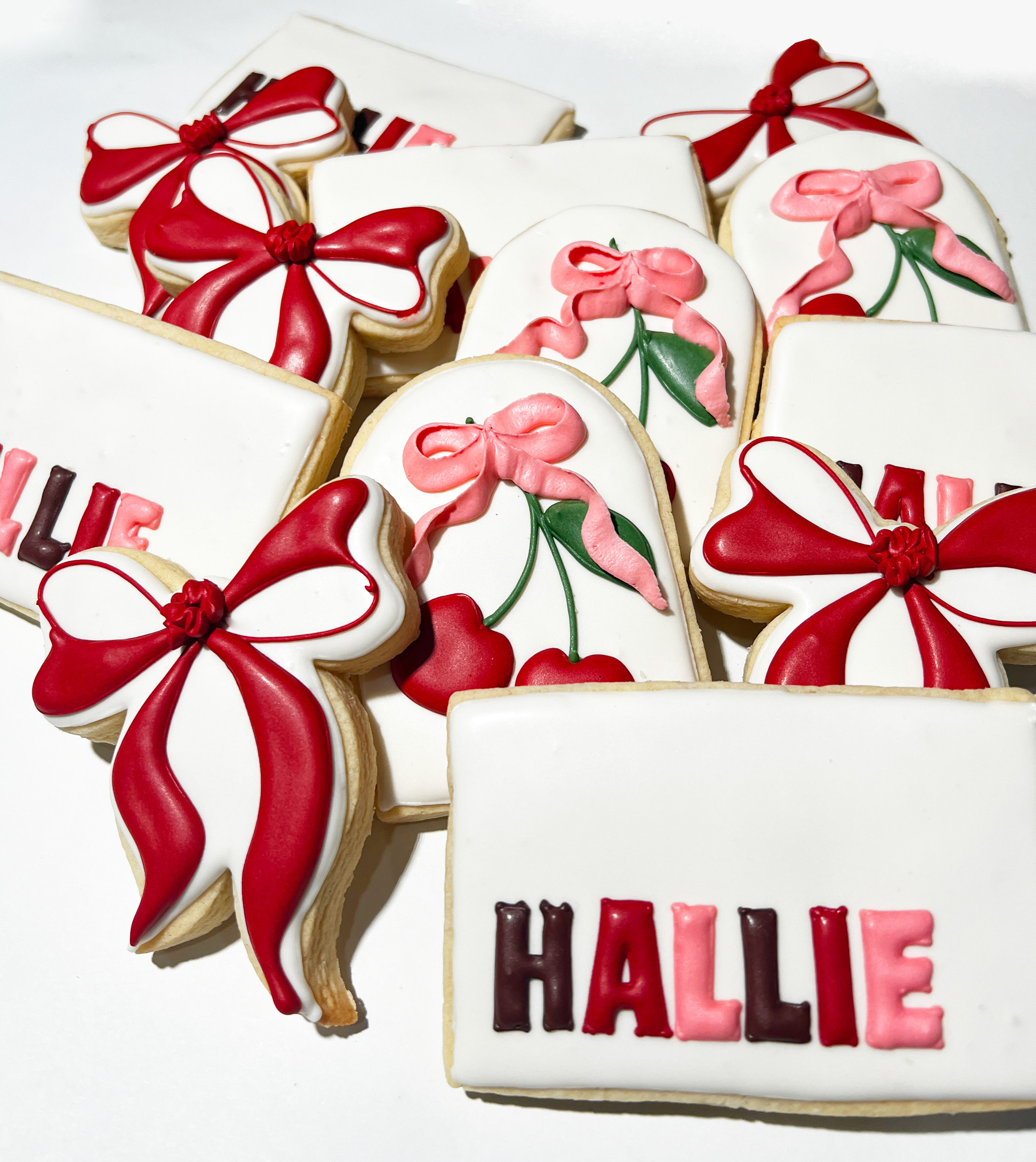 Playful stack of hand-decorated sugar cookies on a white background: white-iced shapes decorated with bold red and pink bows, cherry motifs, and a rectangular cookie with pink-and-red personalized lettering.