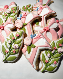 Close-up of hand-decorated sugar cookies with pink ribbon and floral icing, green leaf details on a white background — spring-themed party or shower cookies.