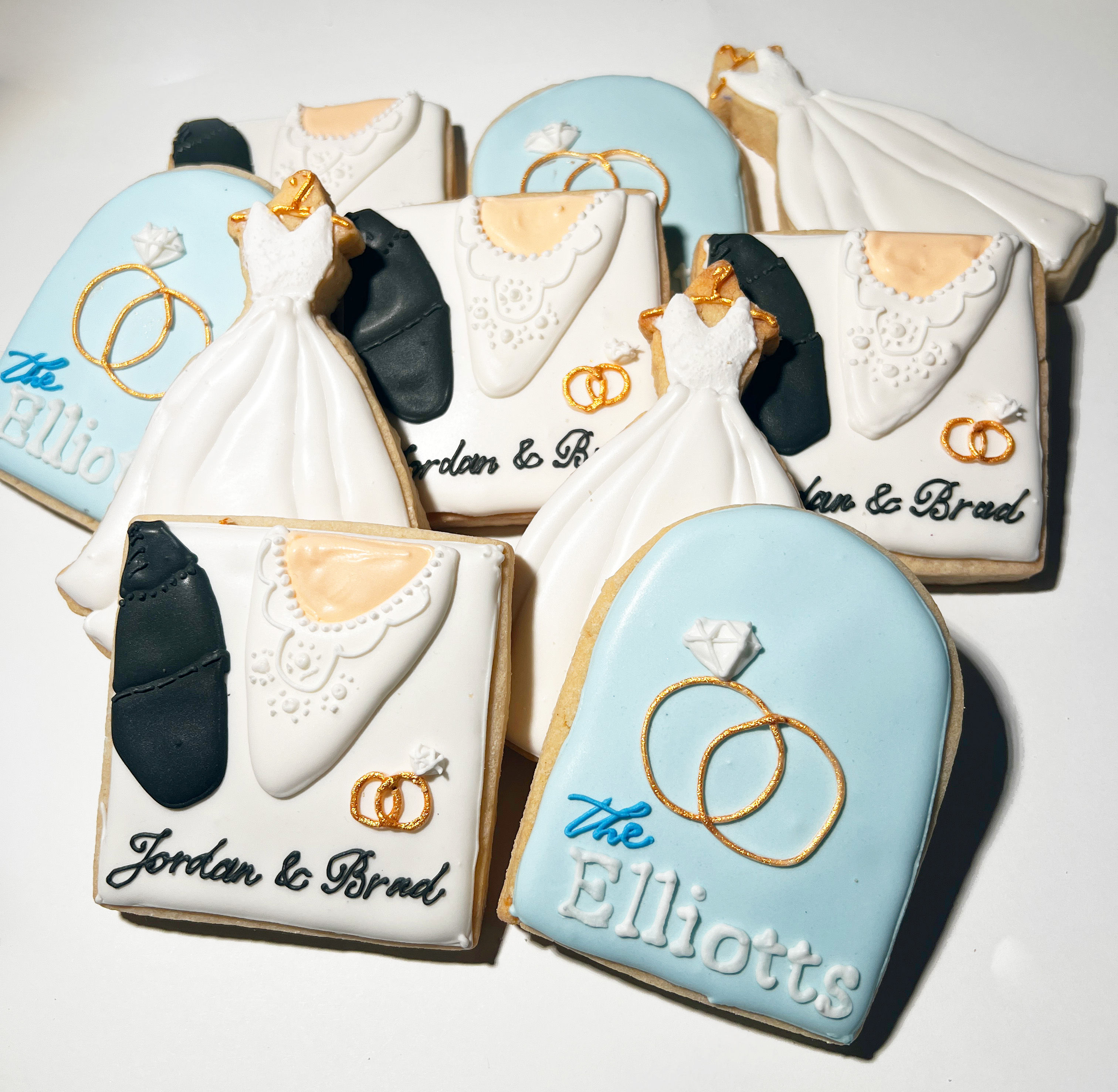 Stack of custom wedding sugar cookies — white iced bridal gowns, tuxedo details, gold ring accents and pale-blue plaque cookies styled as personalized wedding favors.