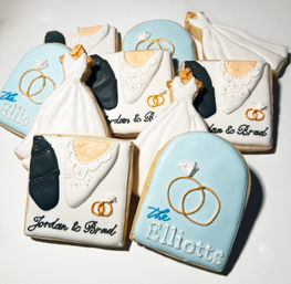 Stack of custom wedding sugar cookies — white iced bridal gowns, tuxedo details, gold ring accents and pale-blue plaque cookies styled as personalized wedding favors.