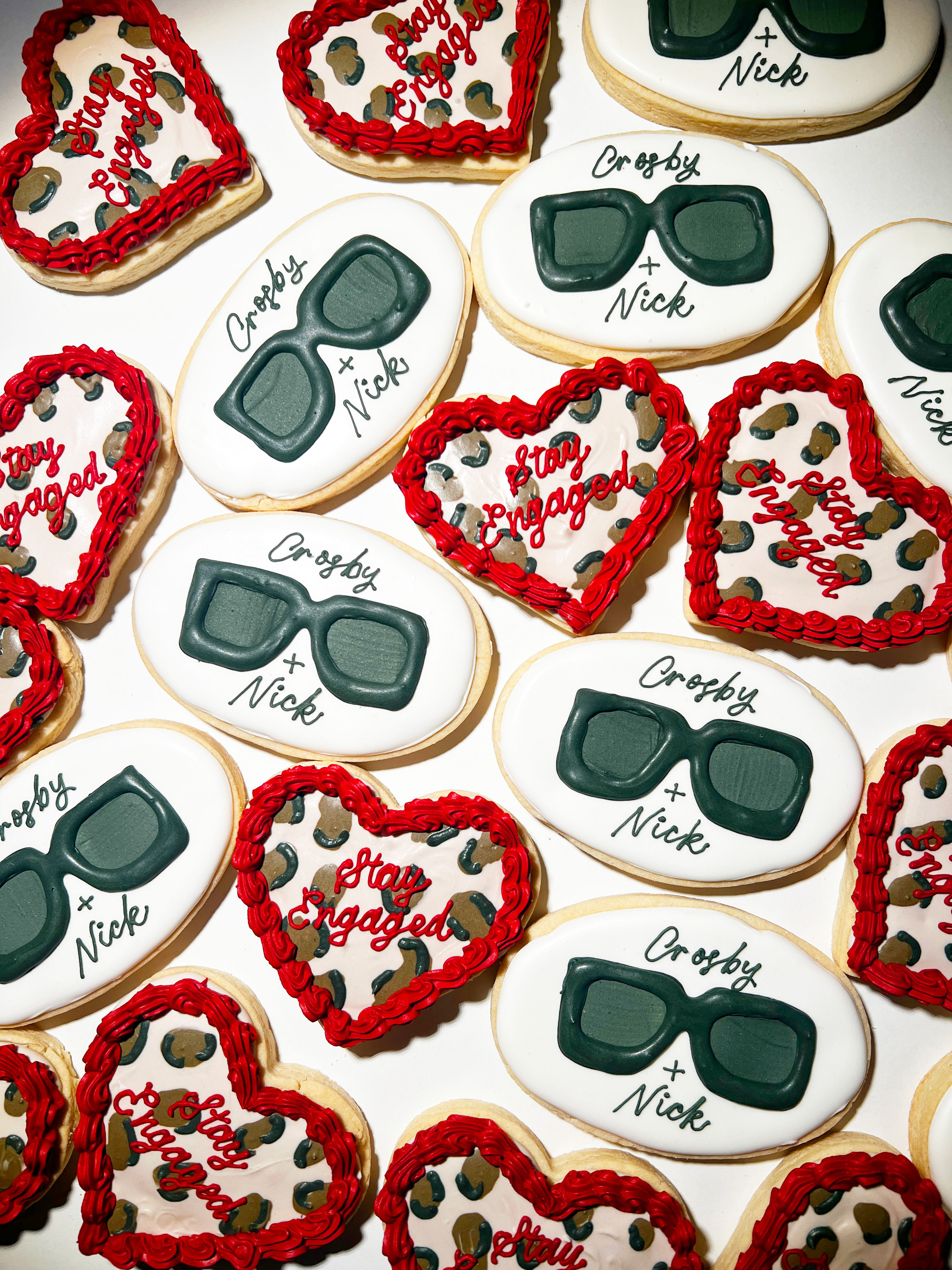 Festive engagement sugar cookies: oval white cookies iced with dark green sunglasses and custom script names, paired with heart-shaped leopard-print cookies edged in red icing and “Stay Engaged” lettering — party-ready dessert assortment.