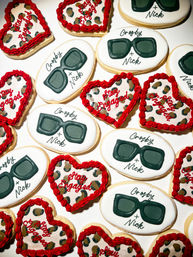 Festive engagement sugar cookies: oval white cookies iced with dark green sunglasses and custom script names, paired with heart-shaped leopard-print cookies edged in red icing and “Stay Engaged” lettering — party-ready dessert assortment.