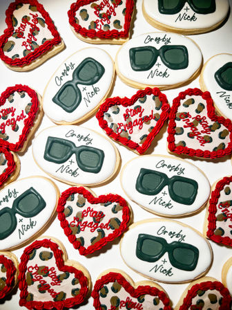 Festive engagement sugar cookies: oval white cookies iced with dark green sunglasses and custom script names, paired with heart-shaped leopard-print cookies edged in red icing and “Stay Engaged” lettering — party-ready dessert assortment.
