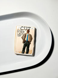 Magazine-cover decorated sugar cookie on a glossy white plate — iced with the word CUT at top and a stylized figure in a brown suit and bright blue tie, flatlay on a minimalist white background.