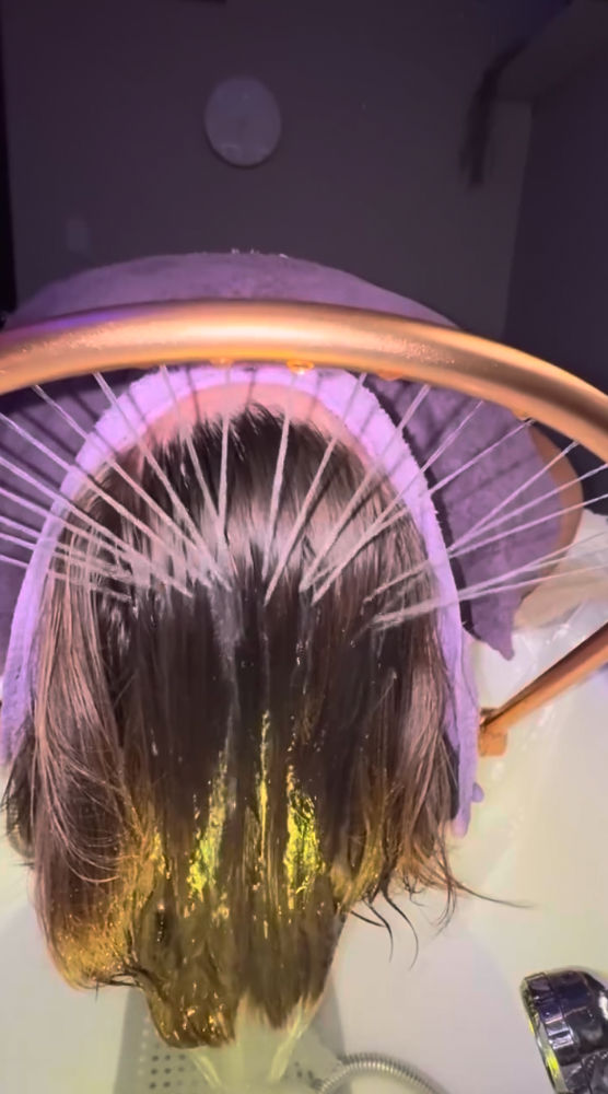 Close-up overhead of wet hair being rinsed into a salon sink under a circular multi-jet shampoo ring, purple towel around the head and warm golden highlights on the strands.