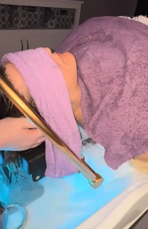 Client reclining at a salon shampoo station with purple towels over face and shoulders, stylist rinsing wet hair over a blue-lit shampoo sink and gold faucet.