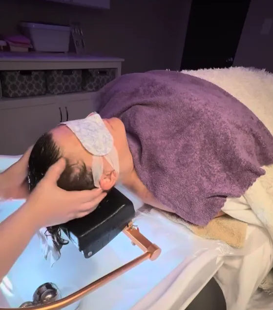 Relaxing salon scalp massage and hair wash — client reclined at sink with eye pads and purple towel.