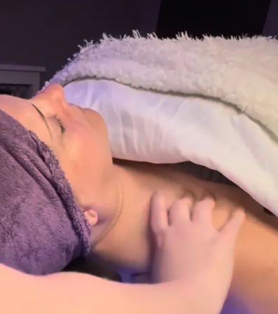 Relaxing spa scene: person with towel-wrapped hair reclined on a spa bed, eyes closed under a plush blanket while a hand gently massages the shoulder and upper chest.