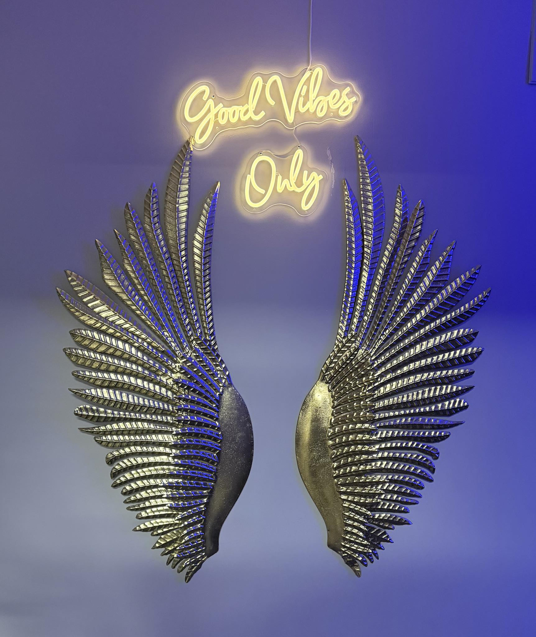 Neon sign reading 'Good Vibes Only' above large metallic angel wings on a purple-blue lit wall — indoor wall decor photo backdrop.