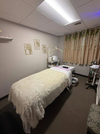 Cozy spa treatment room with plush faux-fur treatment table, neutral curtains with hanging greenery, gold arched lamp and skincare equipment cart.