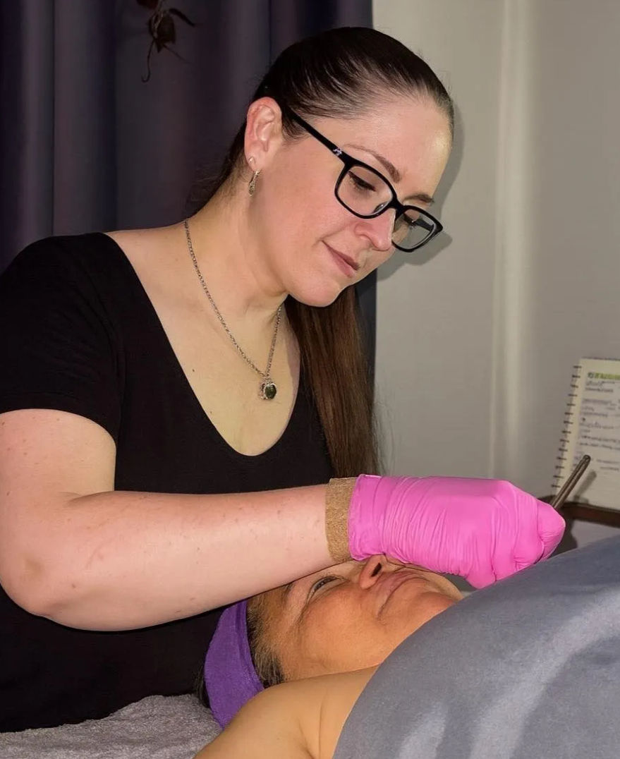 Esthetician in glasses and pink gloves performing a facial treatment on a client wearing a purple headband in a spa treatment room.