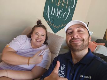 Living room selfie of two smiling adults on a couch giving thumbs-up; woman in light purple T‑shirt and amber bracelet, man in navy polo and white cap, green wall sign and decorative pillows in the background.