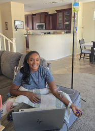 Smiling person on a cozy living room couch working on a laptop while receiving an IV infusion from a standing IV pole, open kitchen visible in the background.