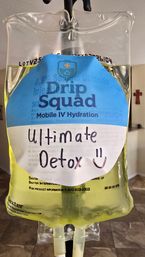 Yellow IV hydration bag with blue-and-white label and handwritten “Ultimate Detox” and smiley, hanging from an IV pole in a home interior