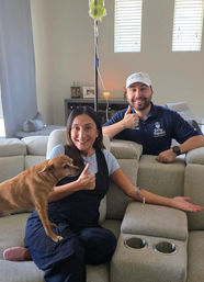 Smiling woman on a living room sofa receiving an at-home IV drip, giving a thumbs-up with a small brown dog on her lap while a uniformed technician in a cap poses behind the couch also giving a thumbs-up; IV pole and bright windows in the cozy living room visible.
