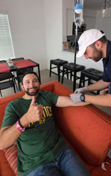 Smiling man gives a thumbs up from an orange couch while a gloved medical technician sets up an at-home IV infusion in a bright apartment living room with dining table and bar stools.