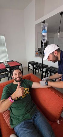 Man on orange couch gives thumbs up while a gloved technician prepares an IV drip for home IV therapy in a modern apartment living room.