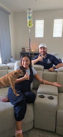 Happy woman receiving at-home IV infusion on a gray sectional couch in a living room, small brown dog on her lap and a smiling healthcare worker in a blue polo giving a thumbs-up beside an IV pole with a yellow-green bag.