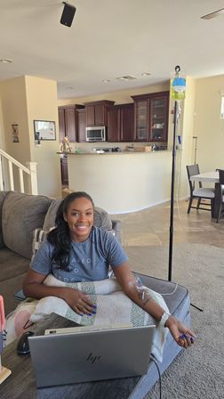 Smiling person relaxing on a living room couch with an IV drip bag on a stand, blanket over their lap and an open laptop on the coffee table, kitchen visible in the background.