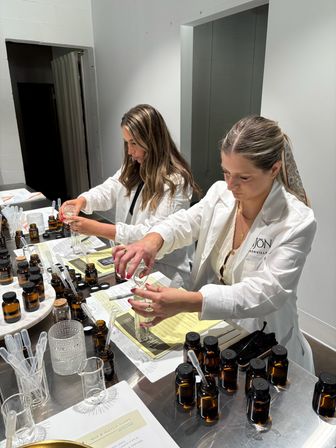 Two people in white lab coats mixing fragrance samples at a scent workshop counter with amber glass bottles, pipettes, beakers, and notes.