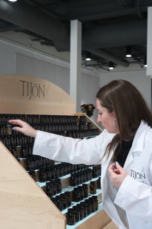 Person in a white lab coat reaches for small amber sample vials on a slanted wooden display rack in a modern fragrance studio.