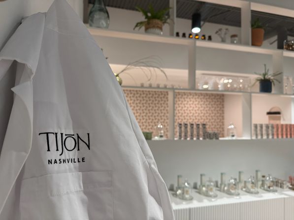 White lab coat with embroidered logo draped in the foreground of a bright, modern fragrance boutique — rows of glass perfume bottles, potted plants, and minimalist white shelving in the background.