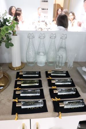 Beauty pop-up tasting table with rows of glass perfume roller bottles on black velvet pouches, clear flip-top water carafes, greenery and a blurred group of people in the background.