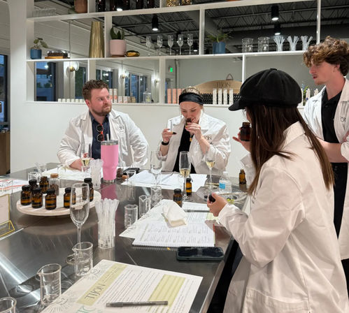Four people in white lab coats sampling amber perfume bottles and essential oils around a stainless-steel table with glassware in a fragrance workshop studio.