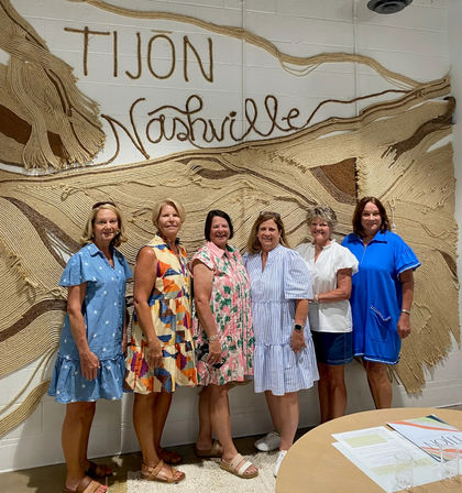 Six women in colorful summer dresses and sandals smiling and posing indoors in front of large textured woven wall art that reads “Nashville”.