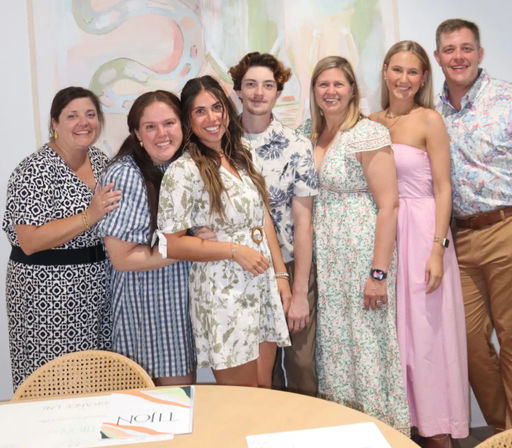 Seven adults smiling and posing in summer dresses and casual shirts in an indoor event space with pastel abstract wall art, standing behind a round table.