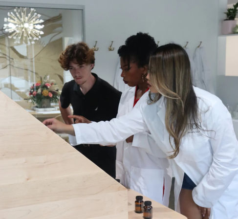 Three people — two in white lab coats and one in a black shirt — examine tiny amber sample bottles on a light wood counter in a modern skincare lab setting with decorative lighting and a floral arrangement.