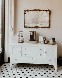 Bright boutique-style coffee station on a white vintage dresser with a single-serve coffee maker, stacked mugs on a tiered stand, tray of stirrers and condiments, ornate framed map on the wall, and black-and-white hex tile floor.